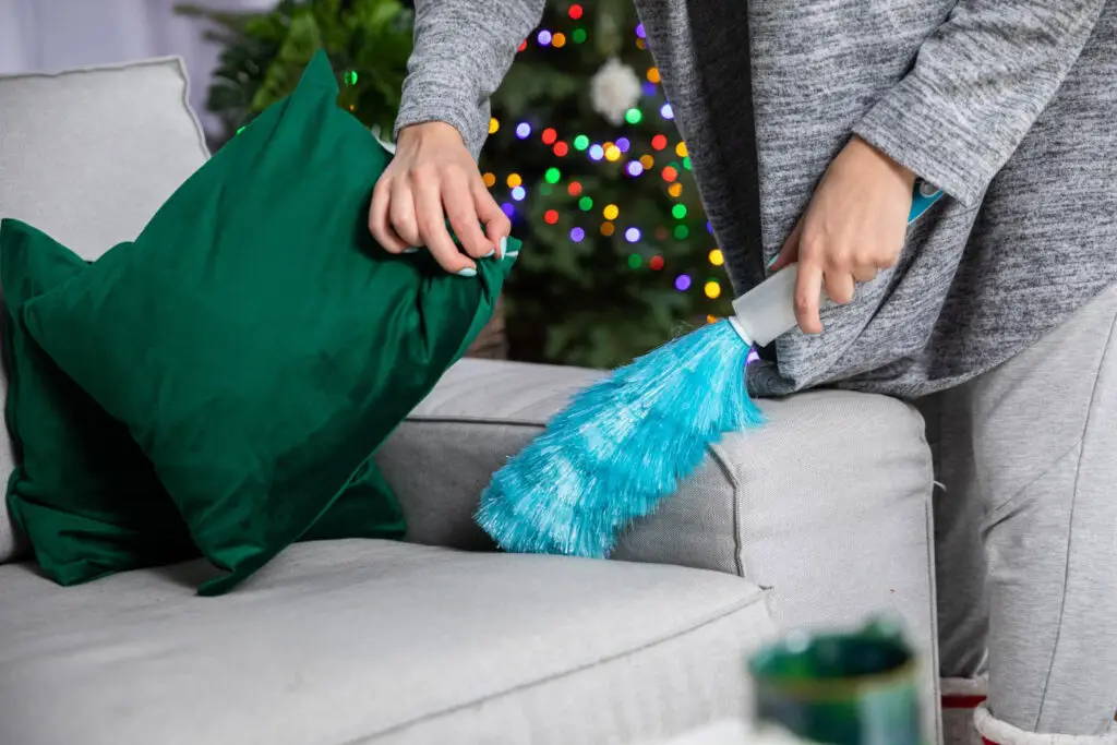 Holiday preparation cleaning: A person dusts a gray sofa with a blue duster, with festive lights visible in the background.