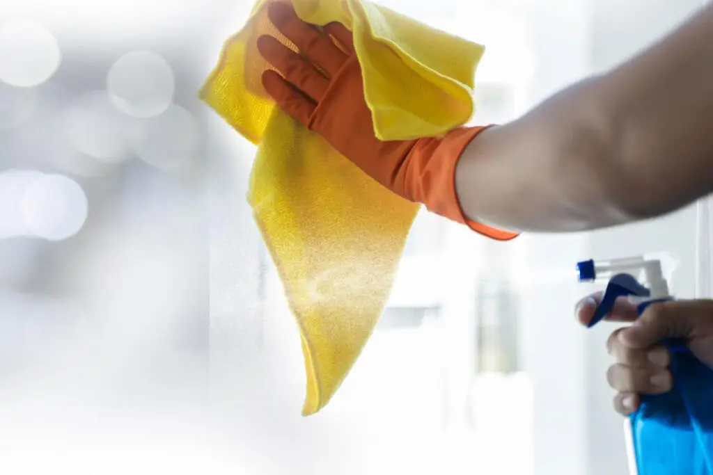 Standard Cleaning: A person in orange gloves sprays and wipes a window with a yellow microfiber cloth.